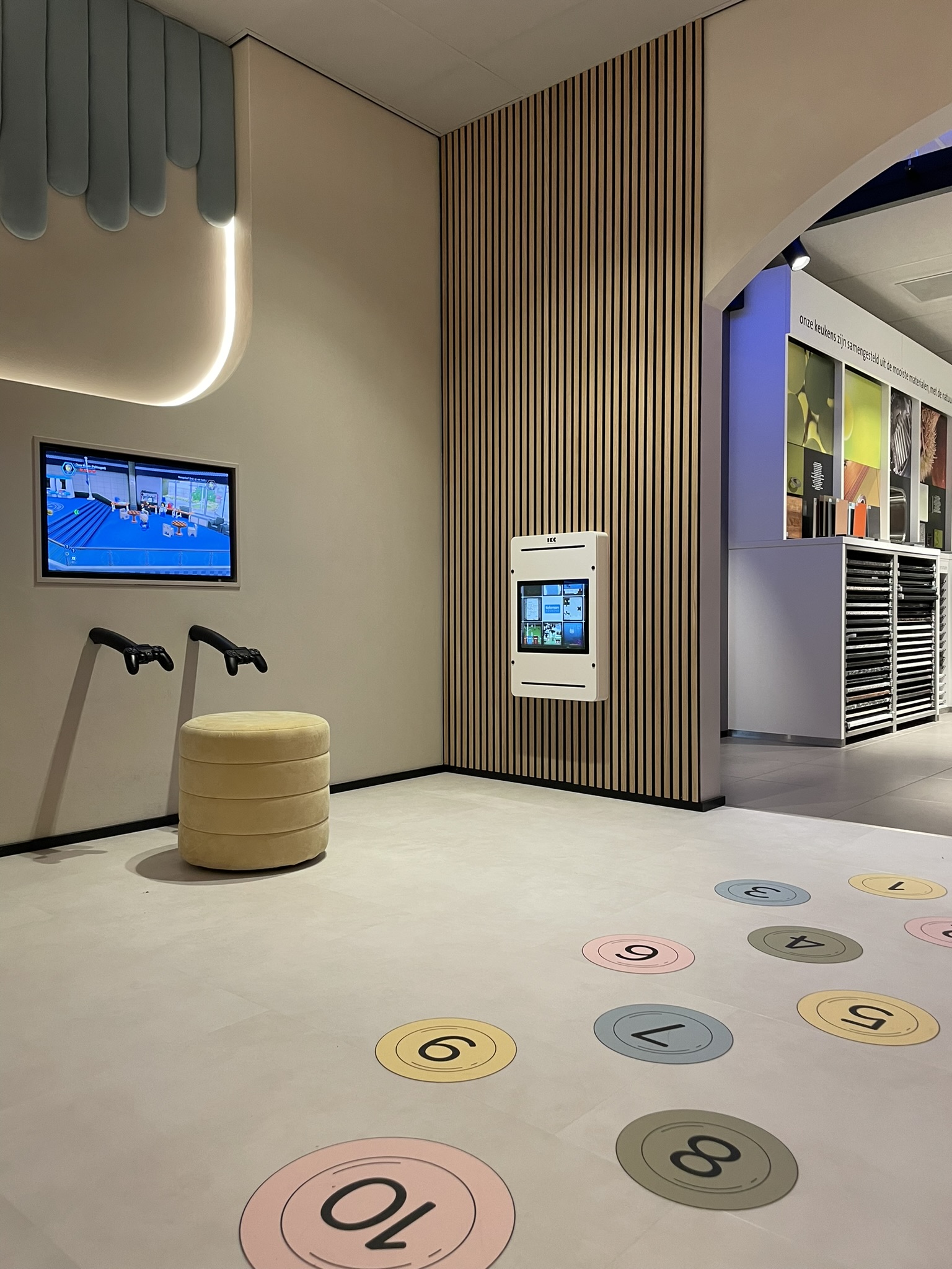 Detail van de interactieve kinderhoek bij Tieleman Keukens, ontworpen door IKC. De foto toont een scherm met gamecontrollers, een ronde zitpoef, een wandtablet en kleurrijke cijfers op de vloer voor een speelse spring- of leerspelactiviteit. De ruimte heeft een rustige, moderne uitstraling met houten lattenwand en zachte wandpanelen.