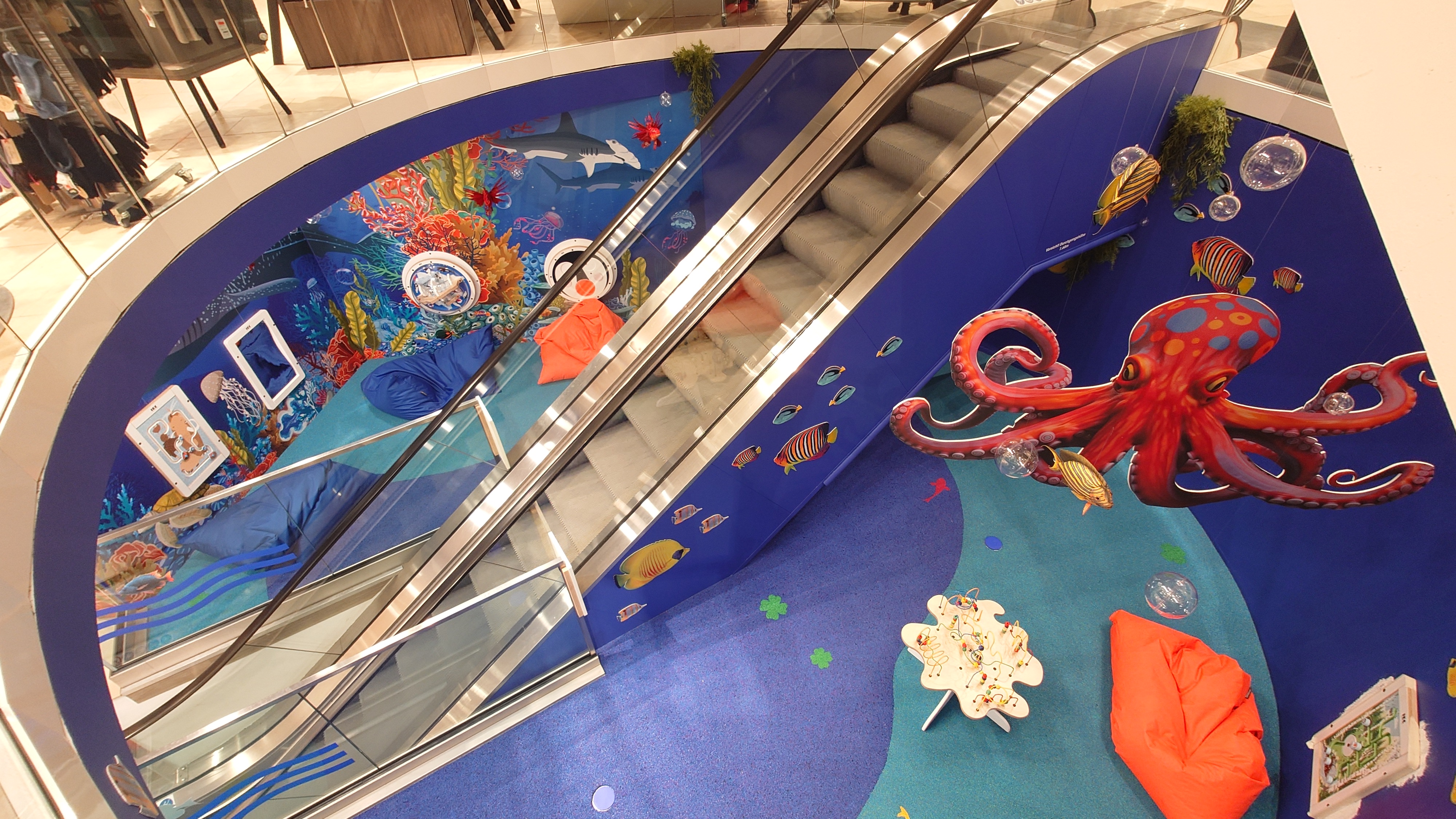 Speelhoek met onderwaterthema bij Modehaus Baltz, ingericht langs de roltrap met kleurrijke muurschilderingen van vissen, een grote octopus, interactieve wandspellen, zitzakken en een speeltafel