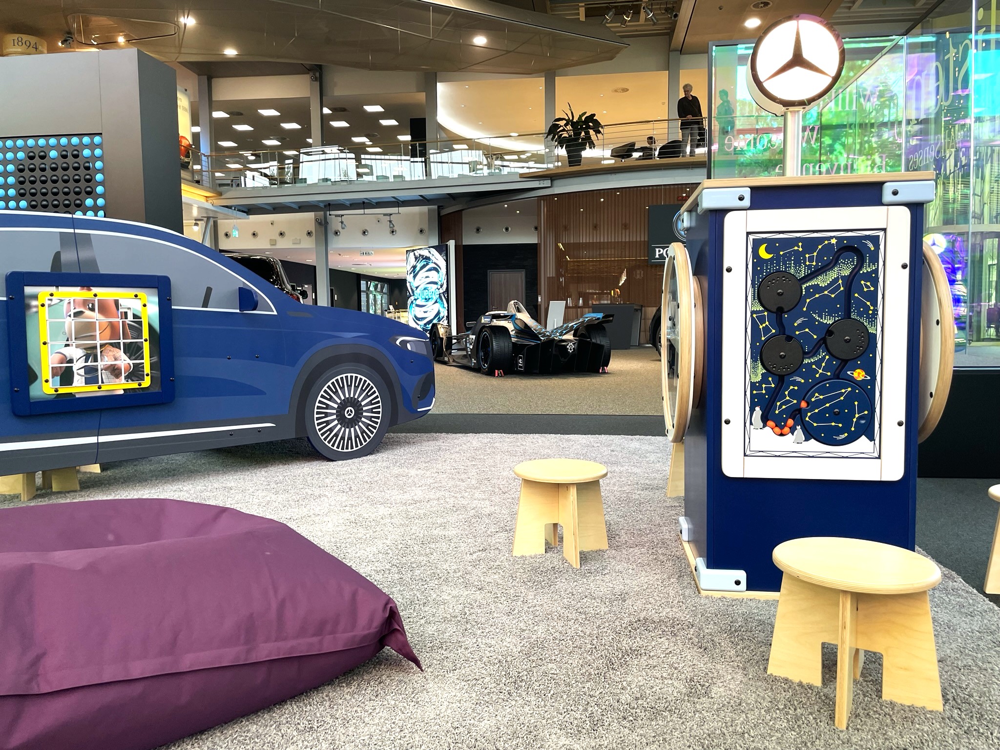 Kinderhoek in Mercedes-Benz Kundencenter met blauw auto-speelmeubel voorzien van wandspel, interactieve speelwand met sterrenhemelthema en Mercedes-logo, houten krukjes en paarse zitzak, in een moderne showroomomgeving.