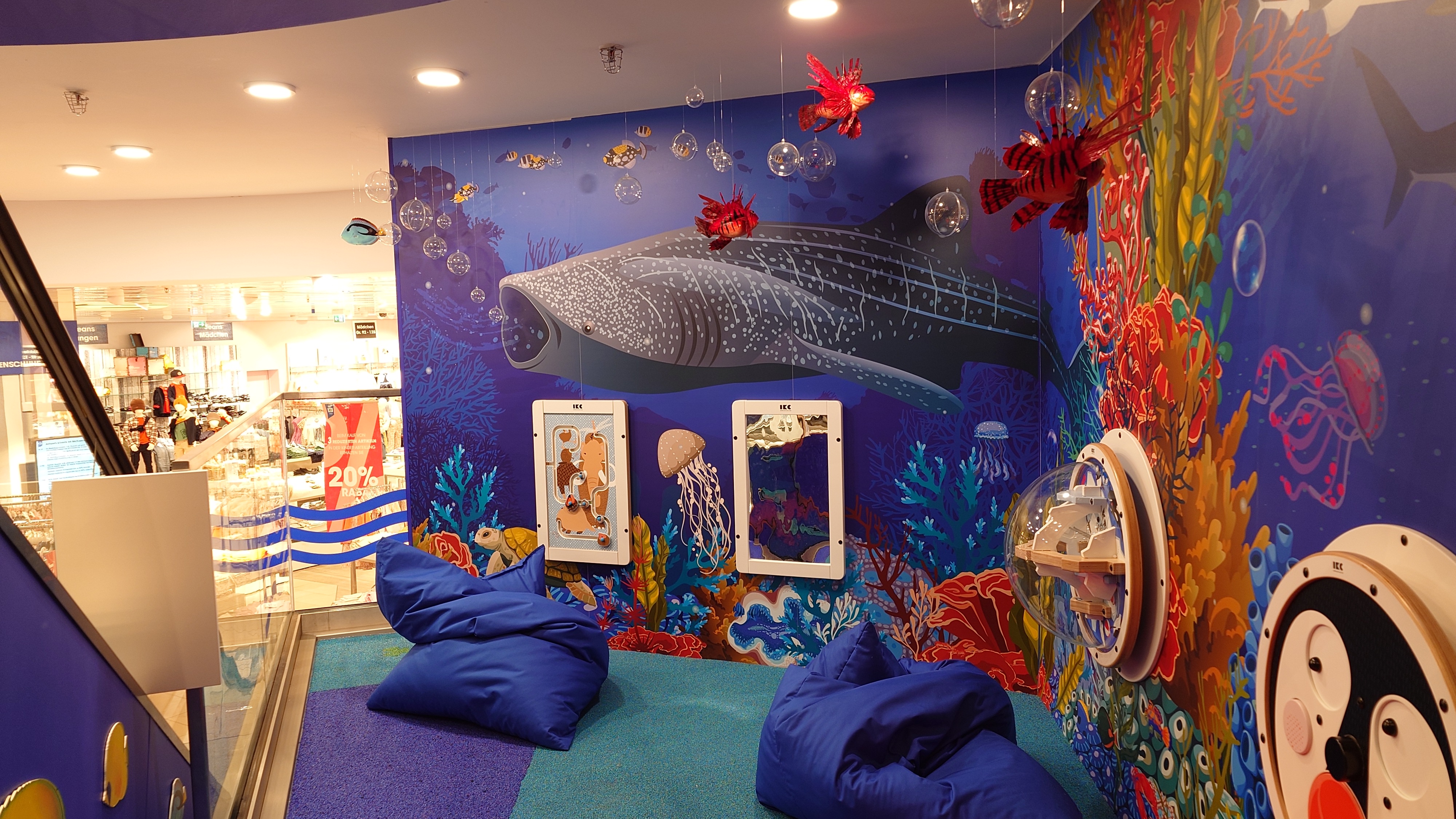 Kinderhoek in oceaanthema met wandspellen, kleurrijke onderwaterillustraties, zitzakken en interactieve speelelementen, gerealiseerd door IKC in een winkelomgeving.