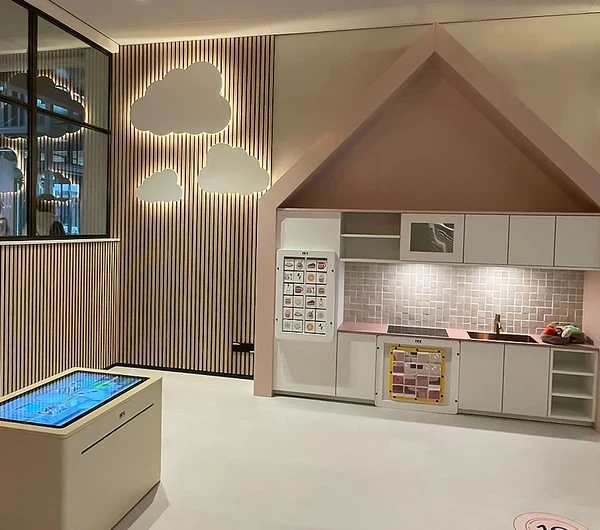 Een sfeervol ingerichte kinderhoek met een speelkeuken in een huisvormig thema, interactieve vloer met hinkelcirkels en een digitale speeltafel. De wand is versierd met houten latten en wolkvormige lichtpanelen. De ruimte is warm verlicht en bevat speelse elementen zoals een houten boomstructuur aan het plafond en retro lampen.