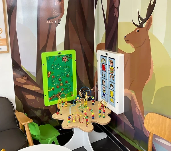 Kinderhoek met wandspelpanelen en kralentafel in een wachtruimte met bosdierenmuurprint. Ontworpen om kinderen op een speelse manier bezig te houden terwijl ouders ontspannen.