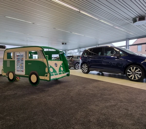 Groene IKC speelbus in de stijl van een klassieke Volkswagen, geplaatst op tapijt in een lichte showroom. Op de achtergrond staan diverse auto's opgesteld; links op de vloer liggen een strandbal en speelgoed.