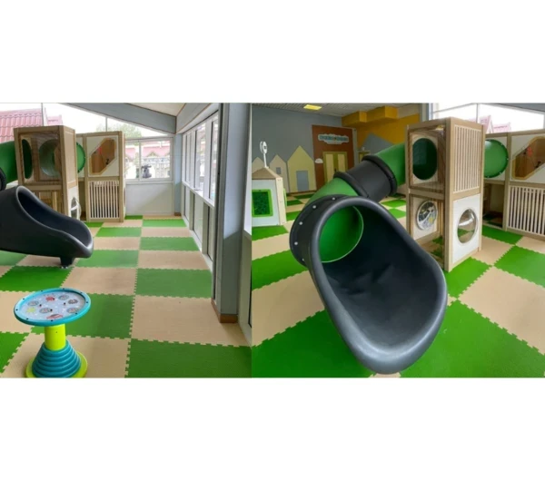 Детский уголок с зелёной горкой, деревянной конструкцией для лазания и игровым столом в светлом помещении с мягким игровым покрытием и большими окнами.