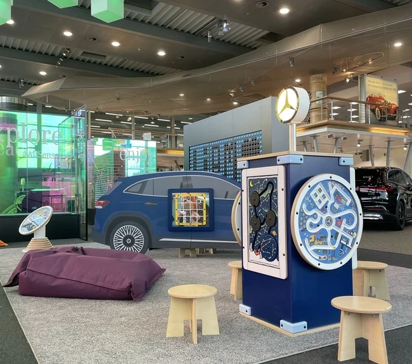 Kinderhoek in Mercedes-Benz Kundencenter met blauw auto-speelmeubel, interactief speelstation met Mercedes-logo, diverse wandspellen, houten krukjes en paarse zitzak, in een kleurrijke en moderne showroomomgeving.