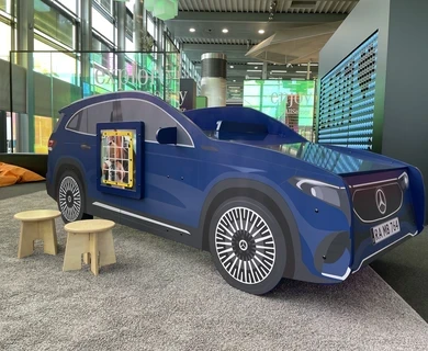 Blauw Mercedes-auto speelmeubel met wandspel, houten krukjes en kleurrijke zitzakken op de achtergrond, geplaatst in de kinderhoek van het Mercedes-Benz Kundencenter in een moderne showroomomgeving.