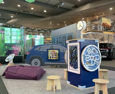 Kinderhoek in Mercedes-Benz Kundencenter met blauw auto-speelmeubel, interactief speelstation met Mercedes-logo, diverse wandspellen, houten krukjes en paarse zitzak, in een kleurrijke en moderne showroomomgeving.