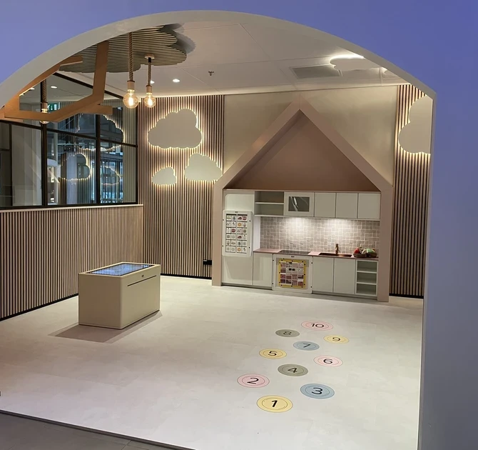 Overzichtsfoto van de kinderhoek bij Tieleman Keukens, ingericht door IKC. De ruimte bevat een speelkeuken in een huisvormige nis, een interactieve speeltafel, wanddecoratie in de vorm van wolken en kleurrijke cijfercirkels op de vloer voor speelse activiteiten. De ruimte is zichtbaar vanuit een boogdoorgang naar de showroom.