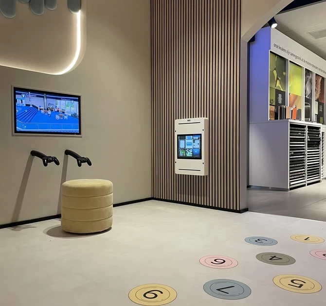 Detail van de interactieve kinderhoek bij Tieleman Keukens, ontworpen door IKC. De foto toont een scherm met gamecontrollers, een ronde zitpoef, een wandtablet en kleurrijke cijfers op de vloer voor een speelse spring- of leerspelactiviteit. De ruimte heeft een rustige, moderne uitstraling met houten lattenwand en zachte wandpanelen.