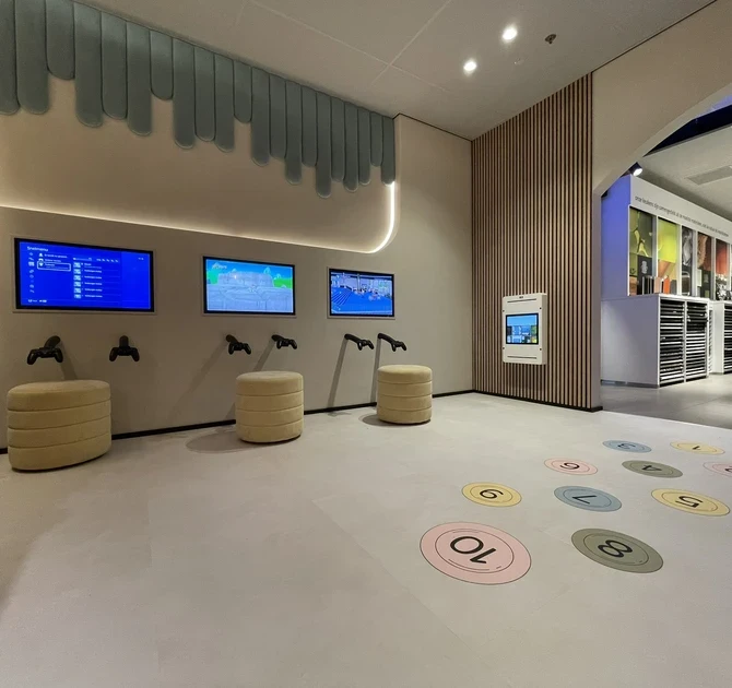 Interactieve gamezone in de kinderhoek bij Tieleman Keukens, ontworpen door IKC. De ruimte bevat drie schermen met gamecontrollers, ronde zitpoefs en speelcirkels op de vloer. De achterwand is sfeervol afgewerkt met een zachtblauwe wanddecoratie en houten latten.