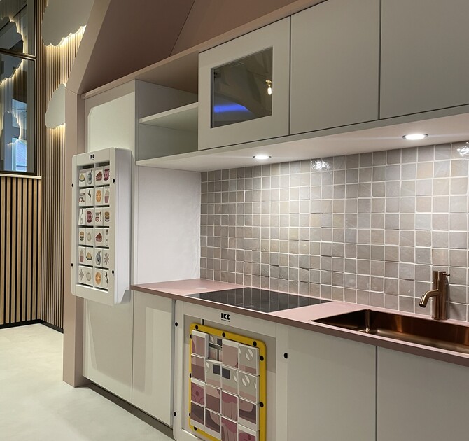 Close-up van de kinderkeuken in de speelruimte bij Tieleman Keukens, ontworpen door IKC. De keuken heeft een roze werkblad, een geïntegreerde kraan en spoelbak, interactieve speelpaneeltjes op de koelkast en oven, en een rustige tegelachterwand. Op de achtergrond zijn wandpanelen en verlichte wolken te zien.