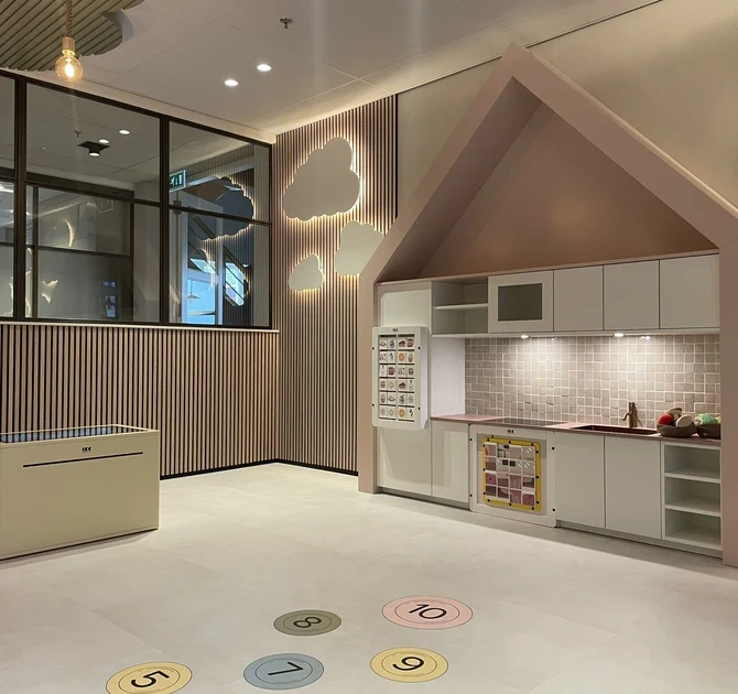 Kinderhoek in de showroom van Tieleman Keukens, ingericht door IKC, met een kindvriendelijke speelkeuken in huisvorm, interactieve speeltafel en speelcirkels op de vloer. De ruimte heeft een warme uitstraling met houten wandpanelen en verlichte wolkdecoratie.