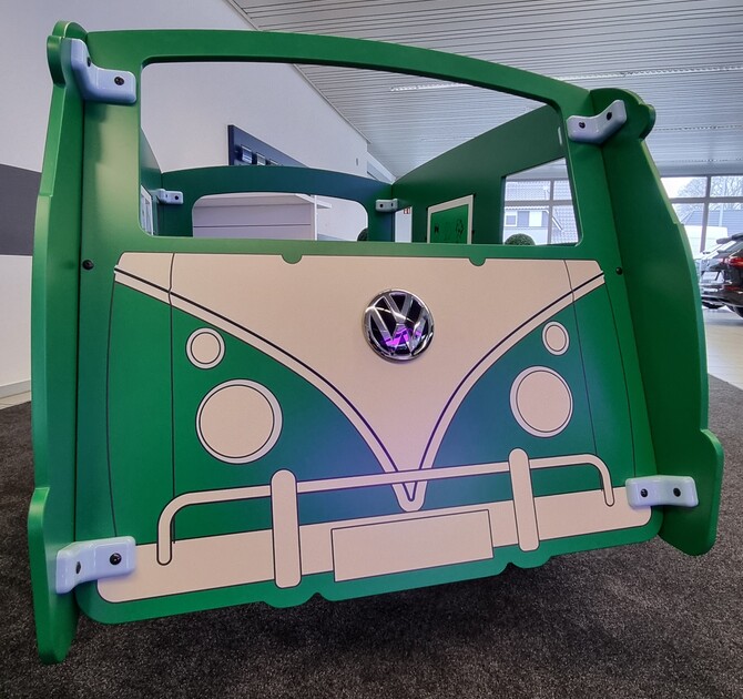 De voorkant van een groene IKC speelbus in Volkswagenstijl, geplaatst in een klantgesprekzone in de showroom van Autohaus Schmidt + Koch. Op de achtergrond een welkomstbord met de tekst "Willkommen bei Volkswagen".