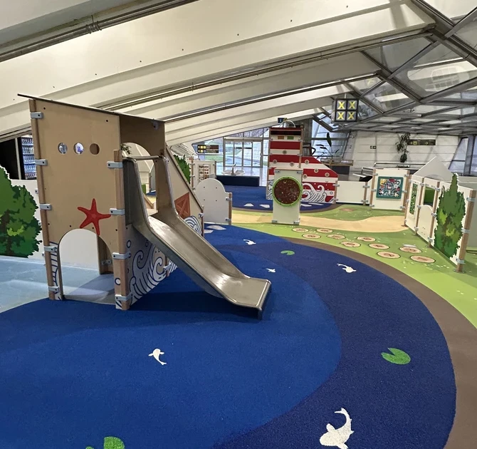 Kinderspeelhoek op Bilbao Airport met blauwe speelkussenvloer, glijbaan in maritiem thema, vuurtoren-speelelement en diverse interactieve spellen in een lichte luchthavenhal.
