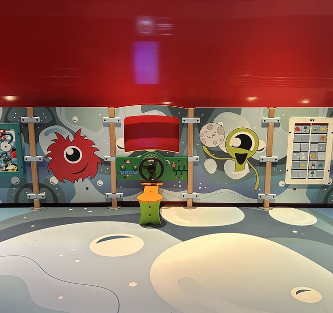 Kinderspeelwand met ruimte-thema, kleurrijke buitenaardse figuren en interactieve spelelementen zoals stuurwiel, schuifpuzzels en spelpanelen, geplaatst tegen een rode wand.