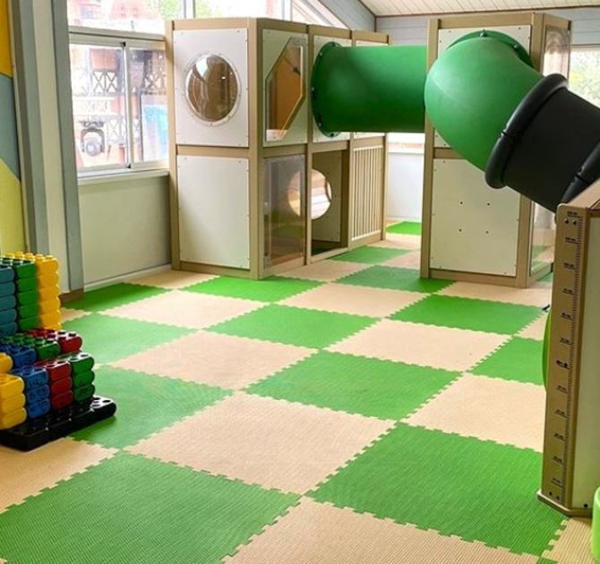 Игровая зона IKC: зелёная винтовая горка, деревянная конструкция для лазания, интерактивный модуль и мягкое покрытие в светлой комнате с большими окнами