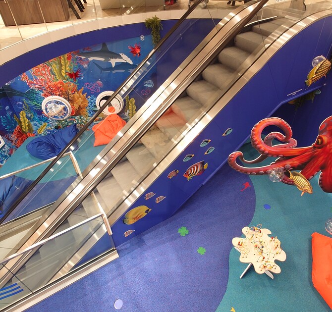 Speelhoek met onderwaterthema bij Modehaus Baltz, ingericht langs de roltrap met kleurrijke muurschilderingen van vissen, een grote octopus, interactieve wandspellen, zitzakken en een speeltafel