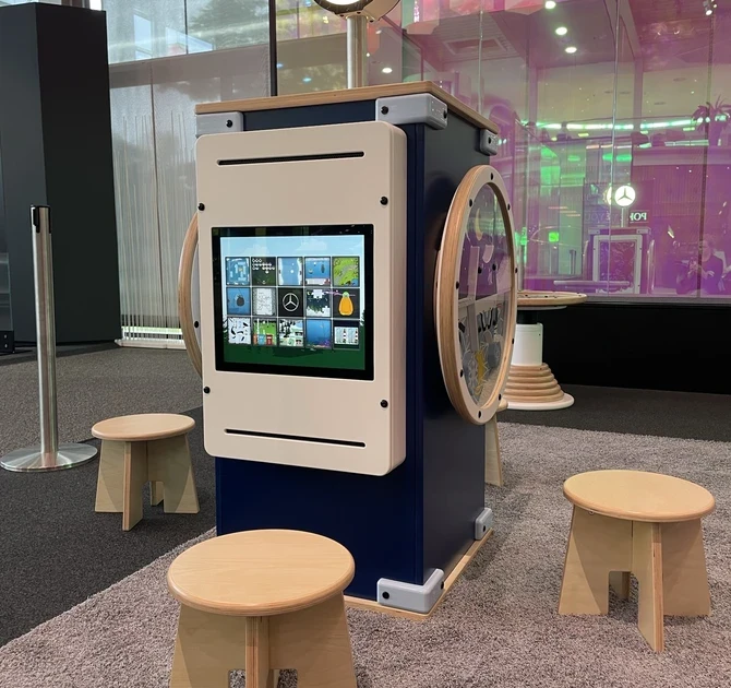Interactief speelmeubel in Mercedes-Benz Kundencenter met digitaal touchscreen vol educatieve spelletjes, houten krukjes en het Mercedes-logo bovenop, geplaatst in een kleurrijke en moderne showroomomgeving.