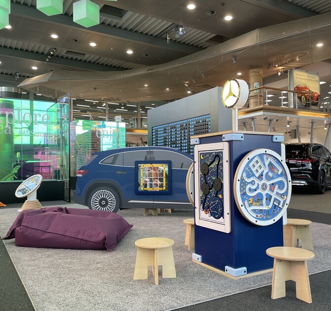 Kinderhoek in Mercedes-Benz Kundencenter met blauw auto-speelmeubel, interactief speelstation met Mercedes-logo, diverse wandspellen, houten krukjes en paarse zitzak, in een kleurrijke en moderne showroomomgeving.