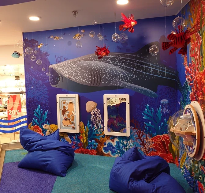 Kinderhoek in oceaanthema met wandspellen, kleurrijke onderwaterillustraties, zitzakken en interactieve speelelementen, gerealiseerd door IKC in een winkelomgeving.